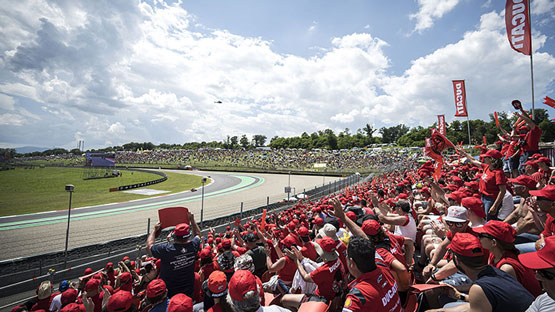 Un grande pubblico entusiasta indossa cappelli rossi, mentre assiste a una gara su un circuito. Il cielo è sereno.