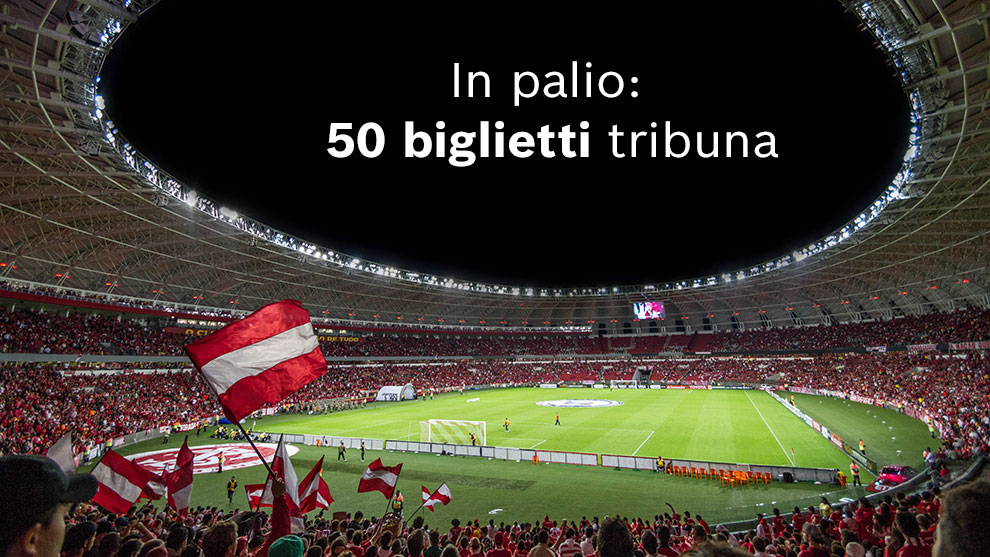 Stadio affollato, tifosi con bandiere, campo da gioco in vista.