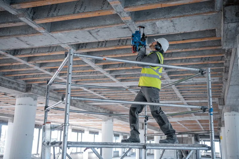 شخص يرتدي معدات السلامة يستخدم مثقابًا أساسيًا على سقف خرساني أثناء وقوفه على السقالة.