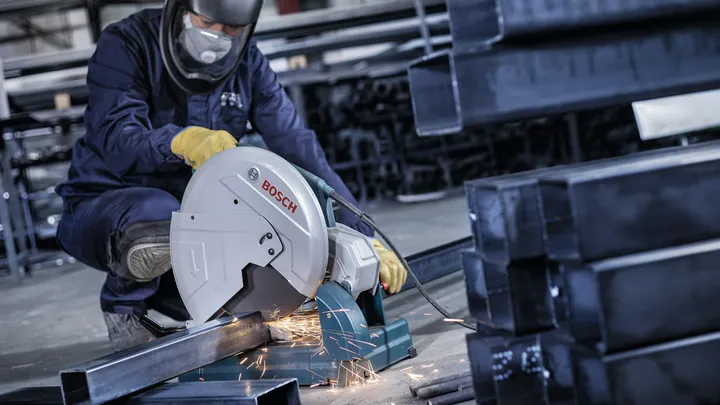 عامل يرتدي معدات السلامة يقطع شعاعًا معدنيًا بمنشار كهربائي، مما ينتج عنه شرارات.