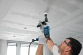 A person wearing safety equipment drills a hole in a ceiling with an impact drill.