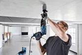 A person wearing safety equipment drills into a ceiling with a dust collection cover attached.