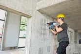 A person wearing safety equipment drills into a concrete wall using a rotary hammer.