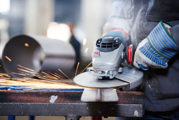 Worker wearing safety equipment grinds metal, producing sparks on a workbench.