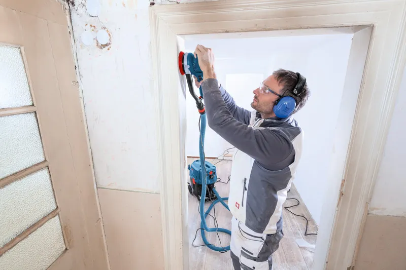 Person wearing safety equipment sands a door frame with a random orbit sander.