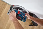 A person operates a cordless circular saw to cut wood flooring near a wall.