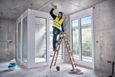 A person wearing safety equipment stands on a ladder using a laser leveling tool indoors.