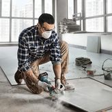 A person wearing safety equipment cuts a tile sheet with a power tool indoors.