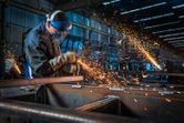 A person wearing safety equipment grinds metal, producing bright sparks in a factory.