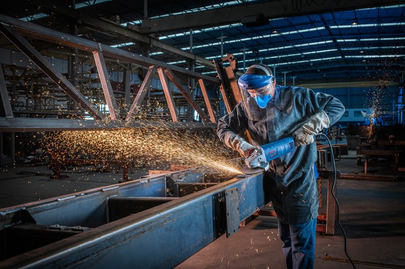 A person wearing safety equipment grinds a metal beam with a large angle grinder.