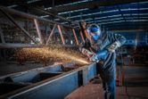 A person wearing safety equipment grinds a metal beam with a large angle grinder.
