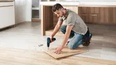 A person drills into a wooden board on the floor, surrounded by wood shavings.