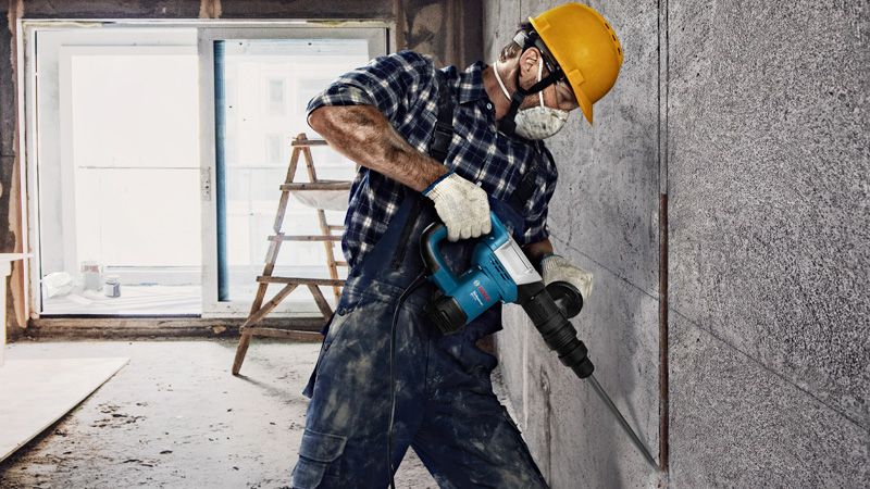 A person wearing safety equipment uses a demolition hammer to break a concrete wall.