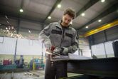 A person wearing safety equipment grinds a steel beam, producing flying sparks.