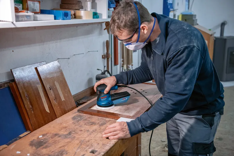 A person wearing safety equipment sands a wooden board with a random orbit sander.