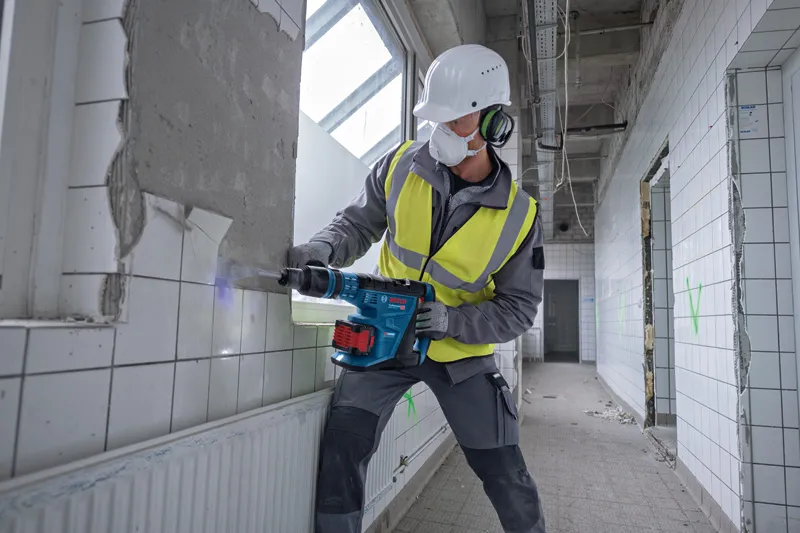 A person wearing safety equipment uses a cordless rotary hammer to break tiled wall.