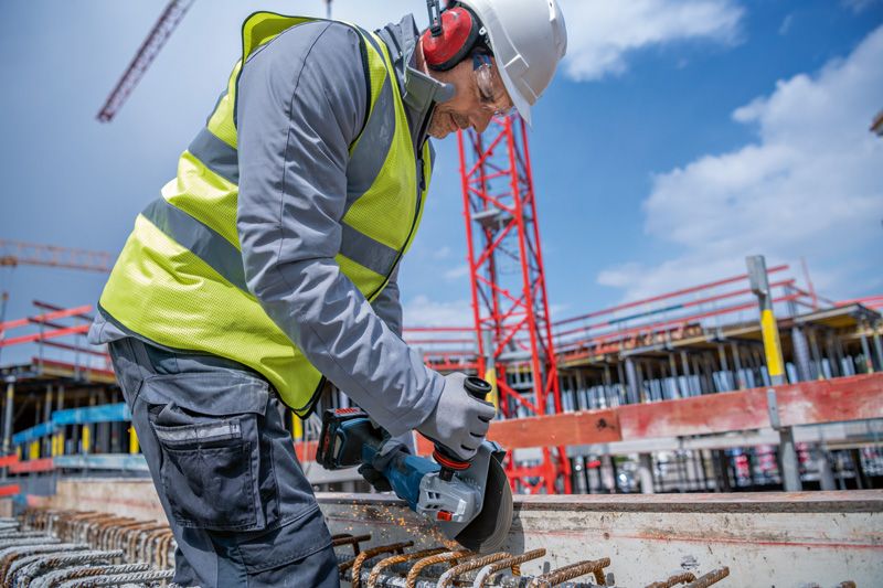 A person wearing safety equipment cuts rebar at a construction site with a cordless angle grinder.