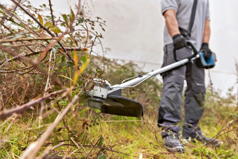 A person wearing safety equipment trims overgrown grass and brush with a cordless brushcutter.