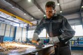 A person wearing safety equipment grinds metal, producing sparks in an industrial workshop.