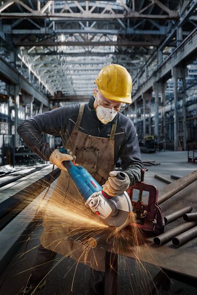 A person wearing safety equipment grinds metal pipes on a workbench in a factory.