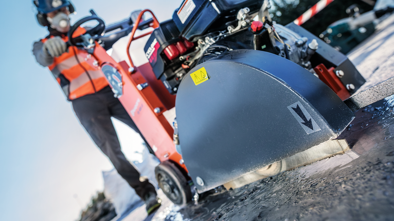 A person wearing safety equipment operates a concrete saw on a wet pavement.