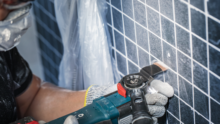 Person wearing safety equipment cuts a hole in wall tiles with a power tool.