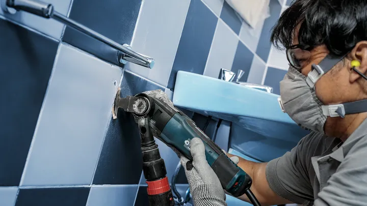 Person wearing safety equipment cuts a hole in a tiled wall with a power tool.