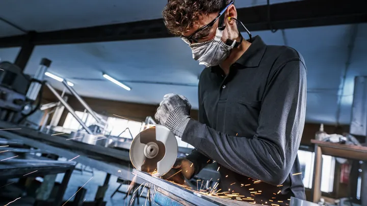 Person wearing safety equipment cuts metal with an angle grinder, sparks flying.