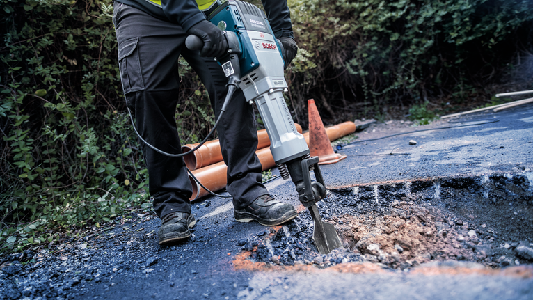 A person wearing safety equipment uses a demolition hammer to break up asphalt.