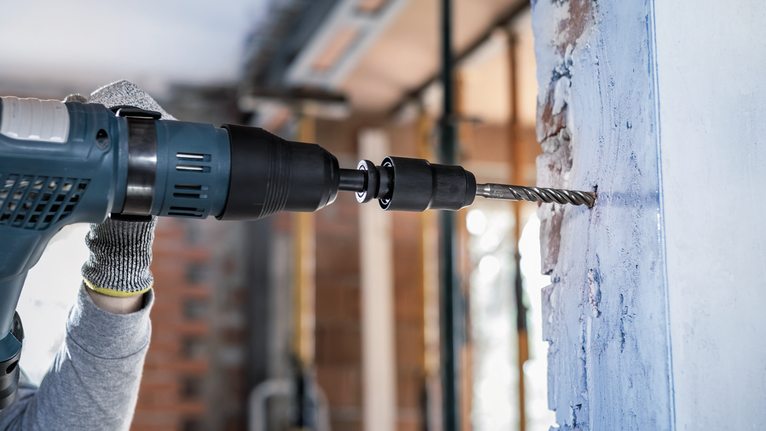 Person wearing safety equipment drills into a concrete wall with a power tool.