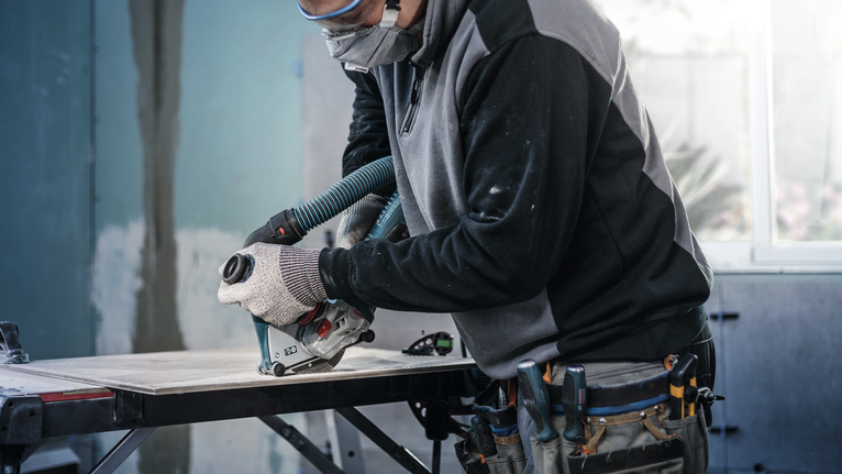 Person wearing safety equipment cuts a tile with a circular saw at a workbench.