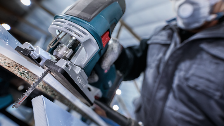 A person wearing safety equipment uses a jigsaw to cut a piece of material.