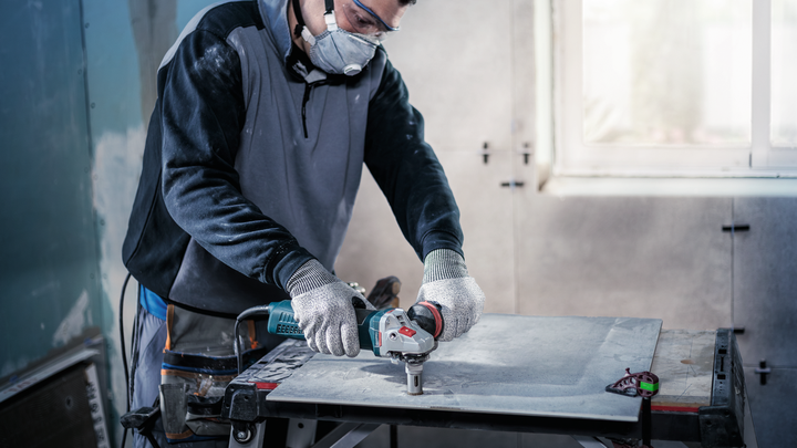 Person wearing safety equipment cuts a tile with an angle grinder indoors.
