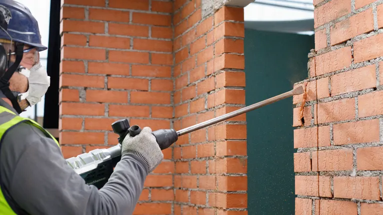 Person wearing safety equipment uses a rotary hammer to chip a brick wall.