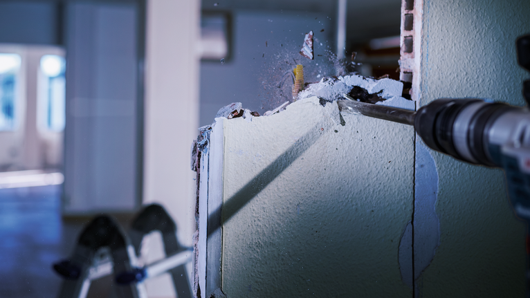 Rotary hammer demolishing an interior drywall, sending debris flying.