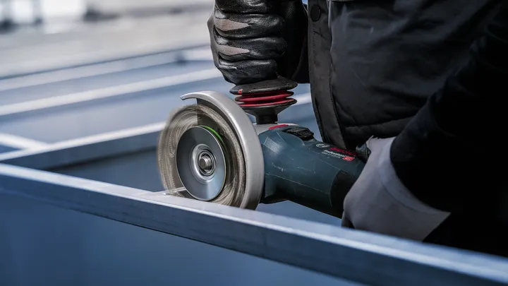 Person wearing safety equipment uses an angle grinder to cut metal beams.