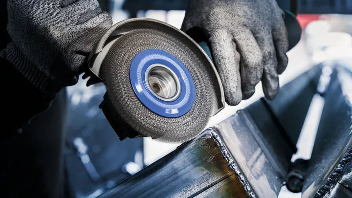 Person wearing safety equipment grinds welded metal beams with an angle grinder.
