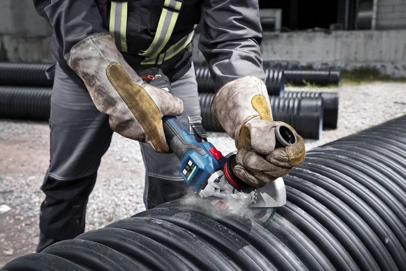 A person wearing safety equipment uses a cordless angle grinder to cut a large plastic pipe.