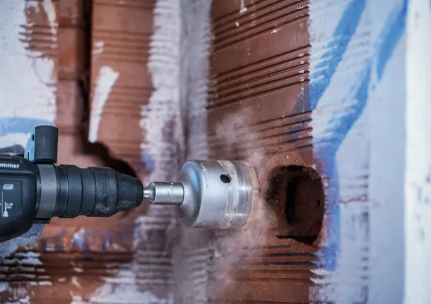 A person wearing safety equipment uses a hole saw to drill into a brick wall.