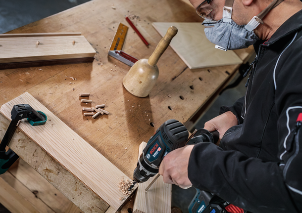 Person wearing safety equipment drills a hole in wooden boards on a workbench.