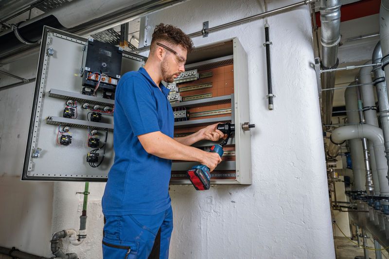 Person wearing safety equipment punches a hole in an electrical cabinet using a cordless tool.
