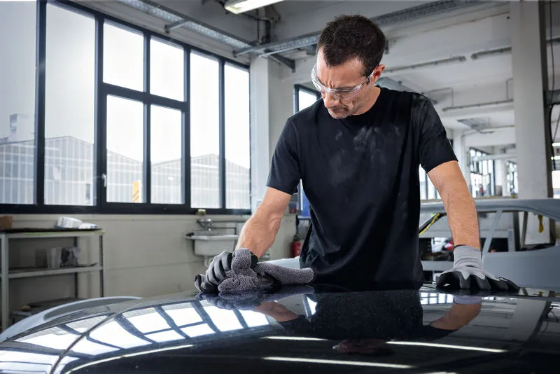 A person wearing safety equipment polishes a car hood with a cloth in a workshop.