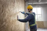 Person wearing safety equipment uses a reciprocating saw to cut into a wooden wall.
