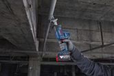 A person wearing safety equipment cuts a metal pipe overhead with a reciprocating saw.