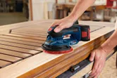 A person sands a wooden door panel with a cordless random orbit sander.