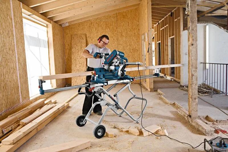 A person wearing safety equipment cuts wood with a miter saw on a wheeled saw stand.