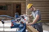 A person wearing safety equipment cuts wood using a mitre saw at a log construction site.