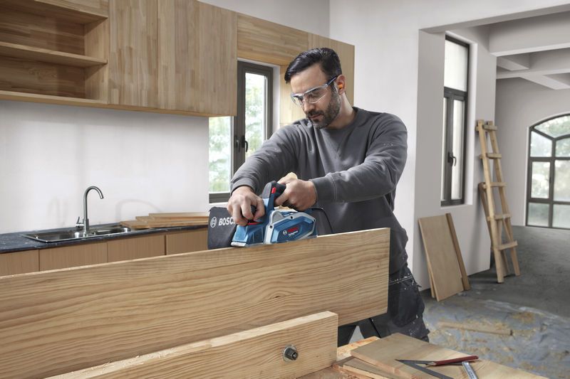 A person wearing safety equipment uses a planer to smooth a wooden board in a workshop.