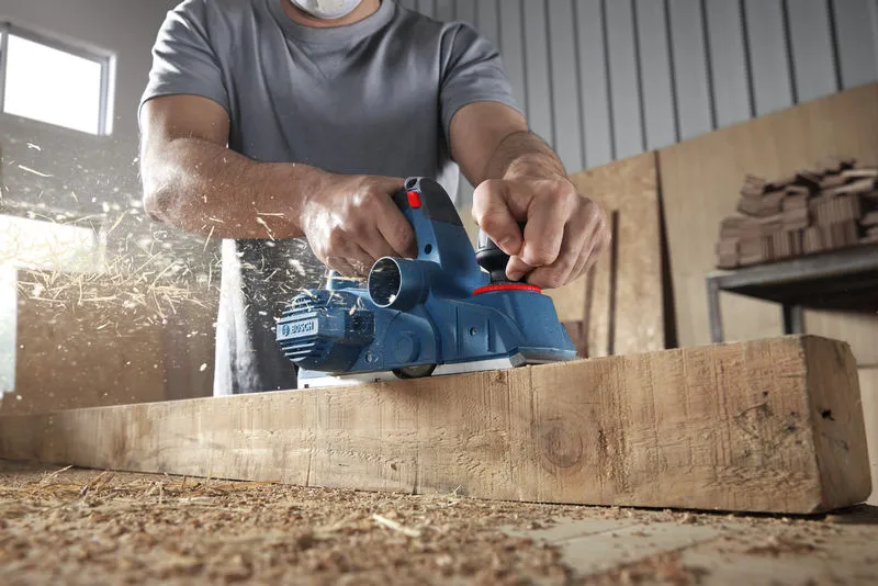 A person wearing safety equipment planes a wooden beam, wood shavings flying.