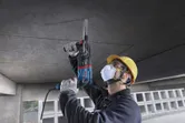 A person wearing safety equipment drills into a concrete ceiling using a rotary hammer.